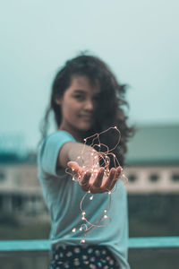 Portrait of young woman holding illuminated string lights while standing against sky