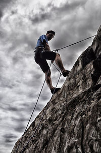 Low angle view of man climbing rock against cloudy sky