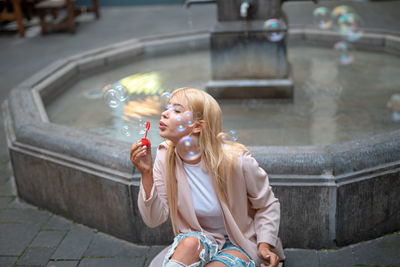 Young woman blowing bubbles while sitting by fountain on footpath