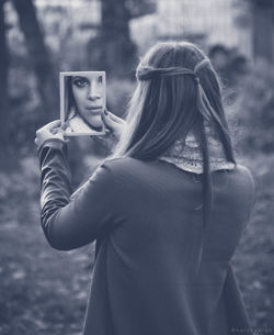 Rear view of young woman holding mirror with reflection in forest