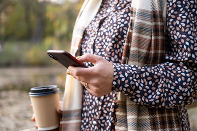 Close up women holding coffee cup, using phone in nature in autumn park in fall