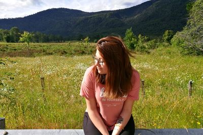 Young woman sitting on railing by grassy field against mountain