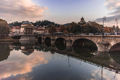 Arch bridge over river by buildings against sky during sunset