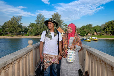 Couple standing on footbridge over river against sky