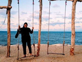 Smiling teenage girl playing on swing at beach against cloudy sky