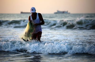 Man holding fishing net while walking in sea against sky during sunset