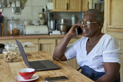 Senior woman sitting at kitchen table using laptop and smartphone