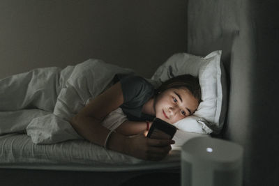 Pre-adolescent girl using smart phone while lying on bed in bedroom at modern home