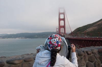 Rear view of girl looking through coin-operated binoculars against golden gate bridge