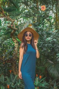 Portrait of young woman wearing sunglasses and hat standing against plants