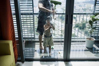 Low section of father with son cleaning windows in balcony seen through glass