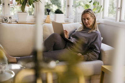 Blond female freelancer with wireless technologies reclining on sofa while working from home