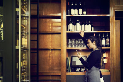 Side view of female owner checking list of wine bottles in phone while standing in cafe