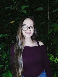 Portrait of smiling young woman wearing eyeglasses standing against plants at night
