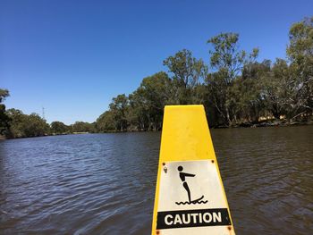 Close-up of yellow sign by lake against clear blue sky