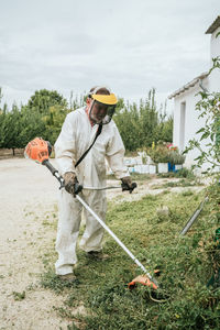 Full body of aged male farmer in white coverall clothes and protective mask cutting grass with weed cutter while working in garden on cloudy summer day