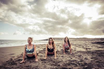 Portrait of friends exercising at beach against cloudy sky
