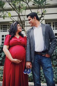 Pregnant couple holding baby booties while standing against plants