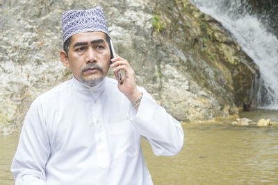 Thoughtful man talking on phone while sitting against waterfall