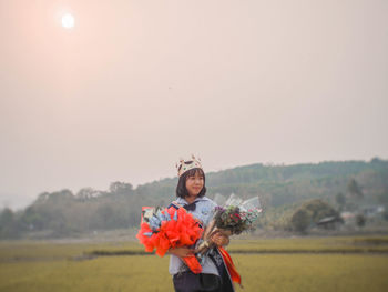 Young woman holding bouquets while standing on field against sky during sunset