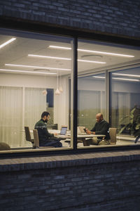 Businessman with male colleague working late in office seen through window at night