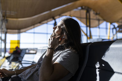 Adult bearded man in stylish outfit with hat sitting in a bench next to glass wall listening to music on wireless earphones while waiting for flight in airport lounge