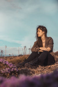 Young woman looking away while sitting on land against sky