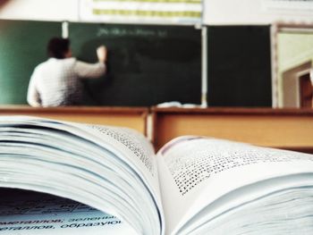 Close-up of open book with teacher writing on blackboard in background