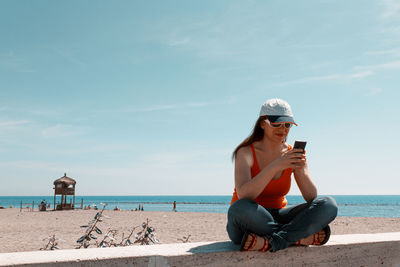 Full length of smiling woman using phone sitting against sea and sky