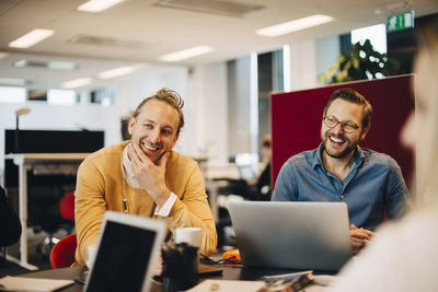 Smiling mid adult male colleagues sitting at conference table while looking away in office