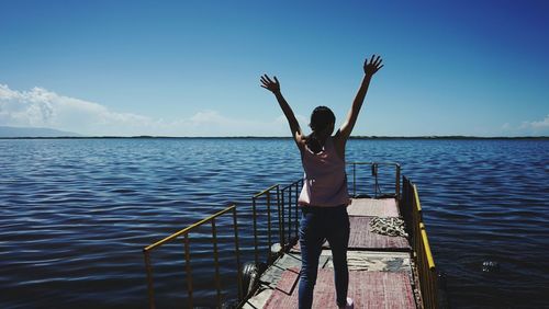 Woman with arms raised walking on pier over sea against sky