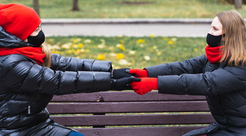 Women wearing mask holding hands against bench at park