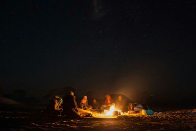 People sitting by bonfire against sky at night