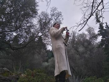 Low angle view of woman photographing on mobile phone while standing against trees