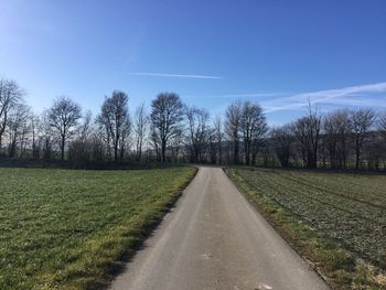 Empty road amidst trees on field against blue sky