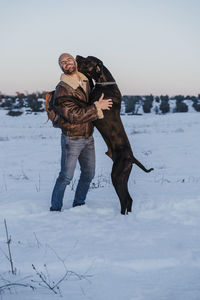 Playful great dane dog leaning on man in snow against clear sky