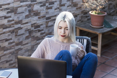 Woman having drink while using on laptop in balcony