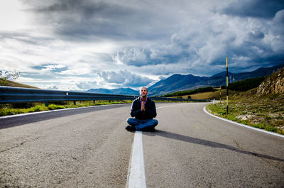 Portrait of man with hands clasped sitting on road against sky