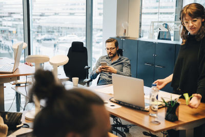 Businessman sitting on chair while using smart phone by businesswoman working in office