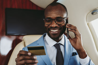 Smiling businessman holding credit card while talking on mobile phone in airplane