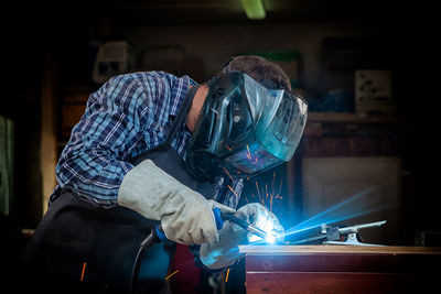 A strong man welder in a black t-shirt, in a welding mask and welders leathers weld metal 