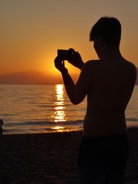 Silhouette man photographing sea against sky during sunset