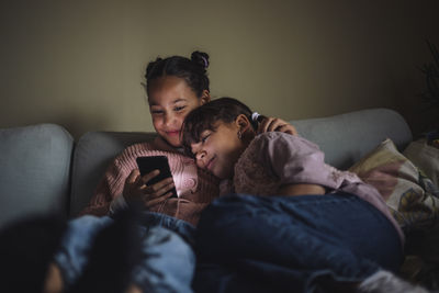 Smiling female siblings sharing smart phone sitting on couch at home