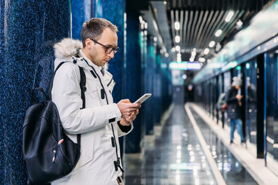 Adult man with wait train at the subway station. male write message or read news on smartphone.