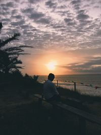 Rear view of man sitting on beach against sky during sunset