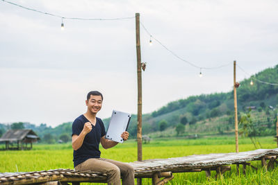 Portrait of man holding laptop while sitting on boardwalk at rice paddy