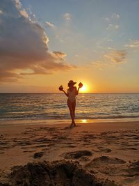 Young woman holding coconuts while standing at beach against sky during sunset