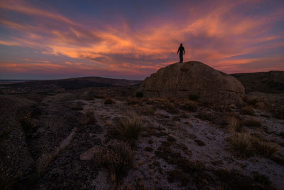 Silhouette of unrecognizable explorer standing on boulder against cloudy sundown sky during trip in mountainous terrain