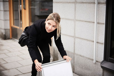 Female real estate agent looking away while arranging blank signboard on sidewalk