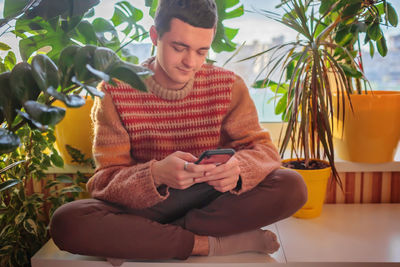 Young man in sweater sitting on table and chatting on smartphone in place among green home plants